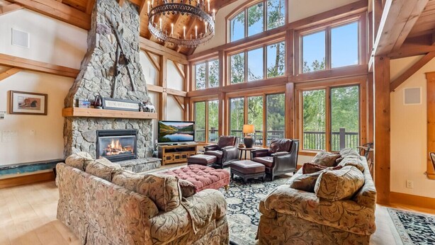 A spacious living room with a stone fireplace, large windows, and wooden beams. - Stunning 5BR Family Lodge—Hot Tub, Huge Deck & Mountain Views (Silverthorne)