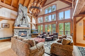 A spacious living room with a stone fireplace, large windows, and wooden beams. - Stunning 5BR Family Lodge—Hot Tub, Huge Deck & Mountain Views (Silverthorne)