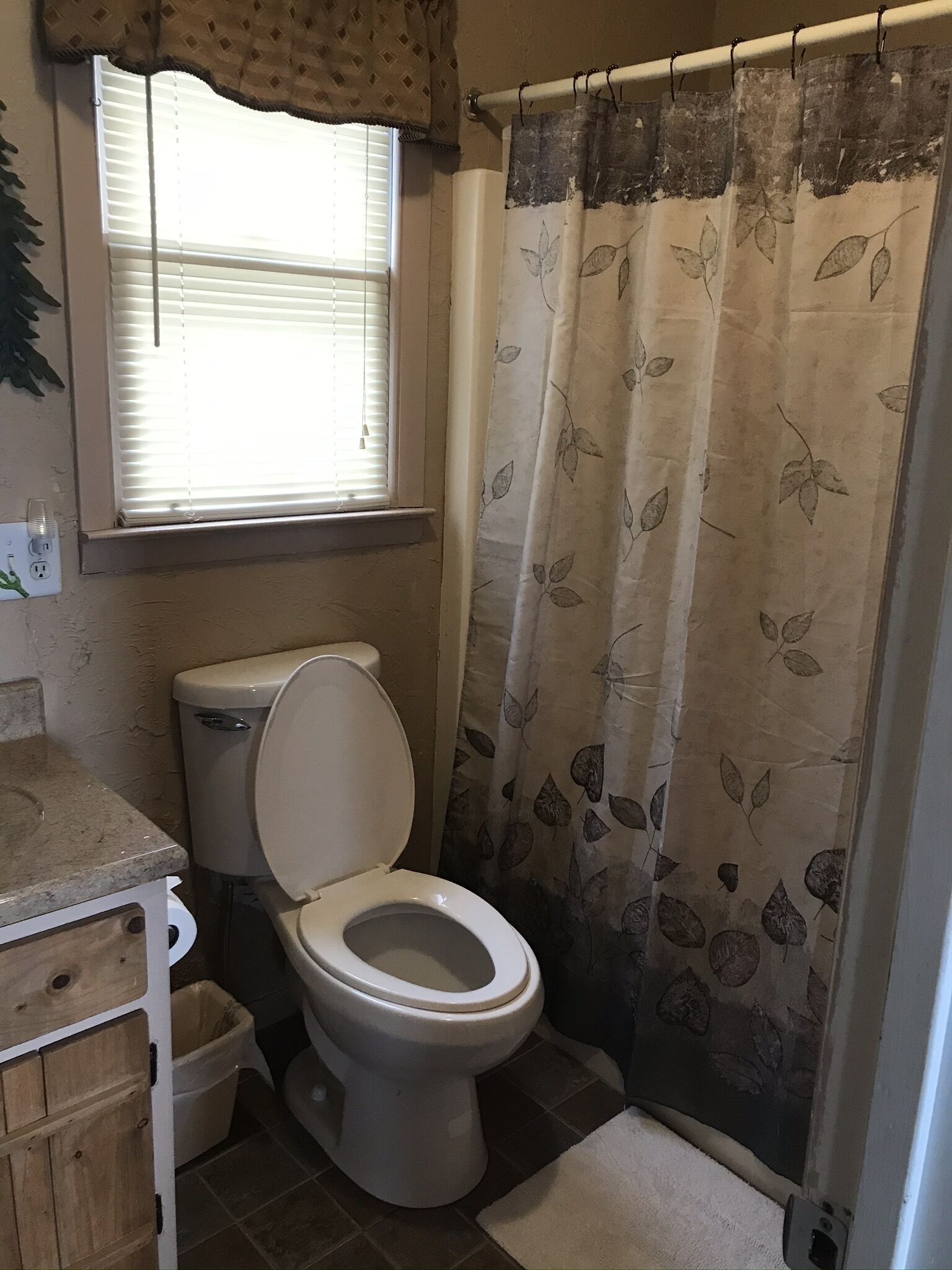 A bathroom with a toilet, a window with blinds, a shower curtain with a leaf pattern, and a tiled floor.