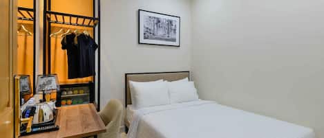 A hotel room with a bed, a desk with a lamp, a chair, and a coat rack with clothes.
