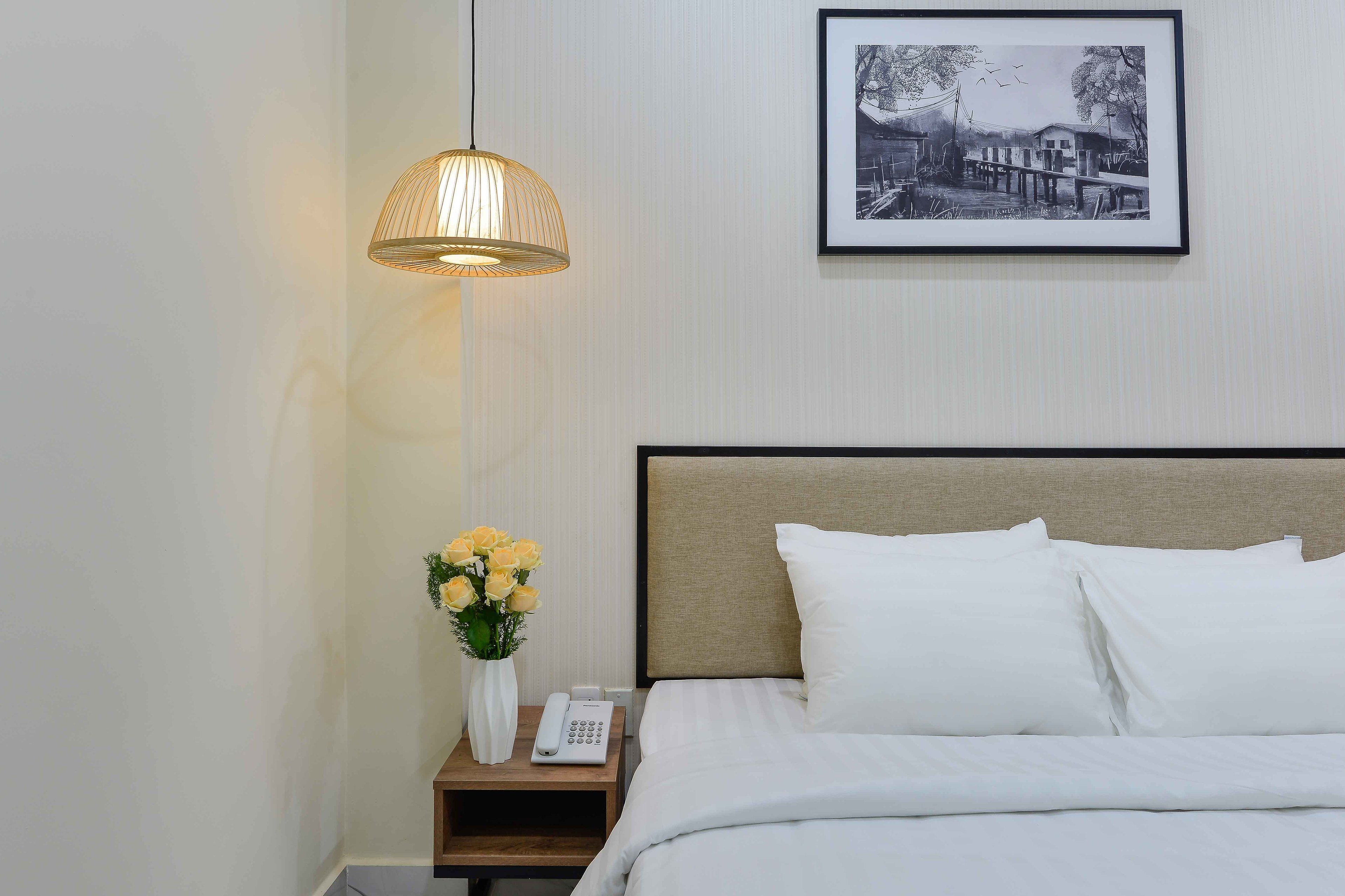 A neatly made bed with white linens, a bedside table with a telephone and a vase of yellow flowers, a hanging lamp, and a framed black and white photograph on the wall.