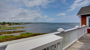 Une terrasse en bois offrant une vue sur une étendue d’eau calme et sur des habitations situées au loin, le long de la côte.