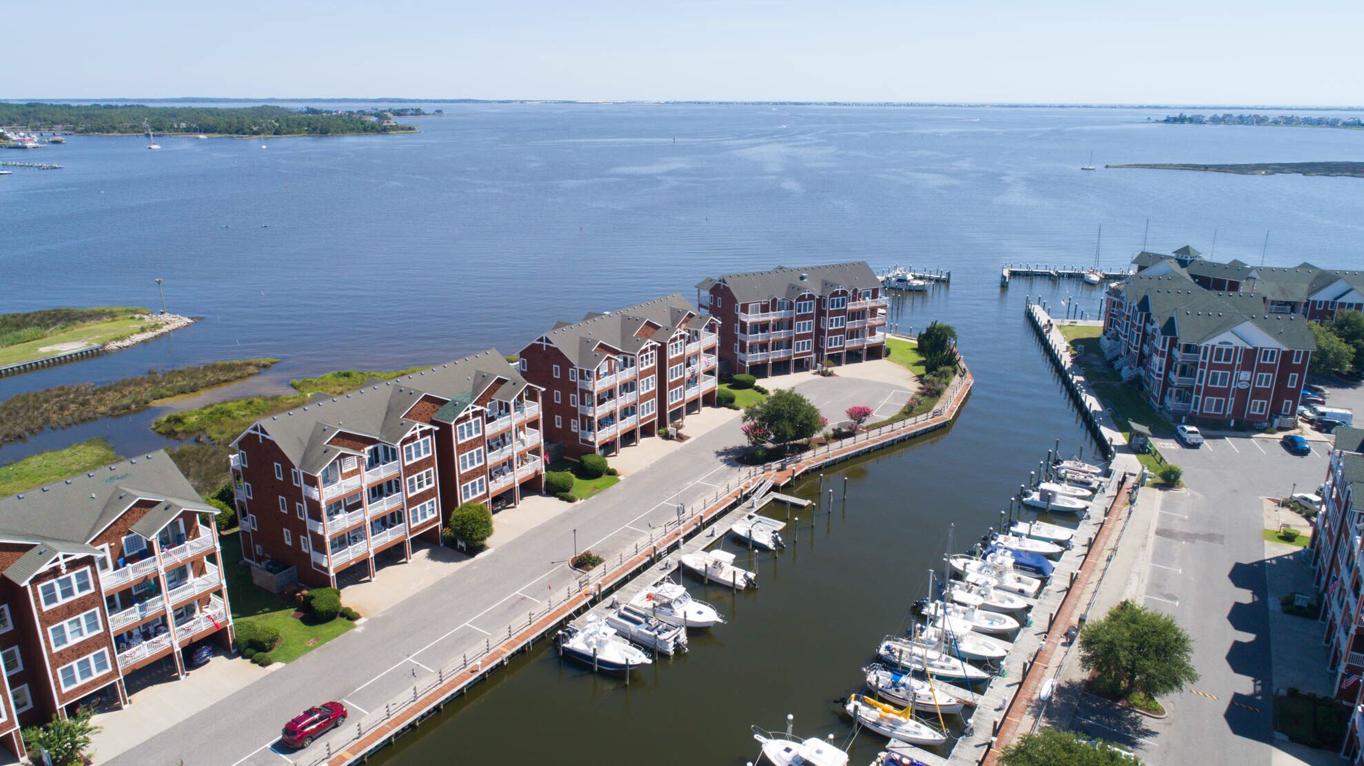A marina with boats docked, surrounded by multi-story buildings with balconies, a road, and a clear view of the water.