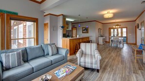 Un vaste salon avec un canapé gris, un fauteuil à rayures et une table basse en bois. On peut apercevoir une cuisine avec des meubles en bois et un coin repas comprenant une table et des chaises.