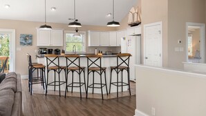 A modern kitchen with white cabinets, a central island with bar stools, pendant lighting, and a view of the outdoors. - Banks Retreat: Private Pool, Hot Tub, Dog-friendly, 700 feet to Beach Access (Corolla)