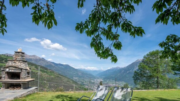 Une cheminée en pierre avec un banc, située dans un espace verdoyant, d’où l’on a une vue sur une vallée et des montagnes.