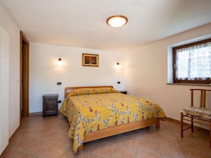 Une chambre à coucher comprenant un lit, une chaise, une table de chevet, une fenêtre avec des rideaux et un tableau accroché au mur.