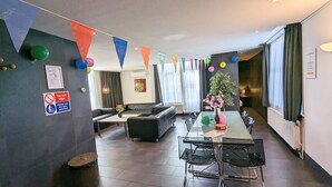 Moderni hotellihuone, jossa on ruokailutila, sohva, pieni pöytä tuoleineen ja ruukkukasvi. Seinillä on värikkäitä, kolmiomaisia lippuja ja ilmapalloja.