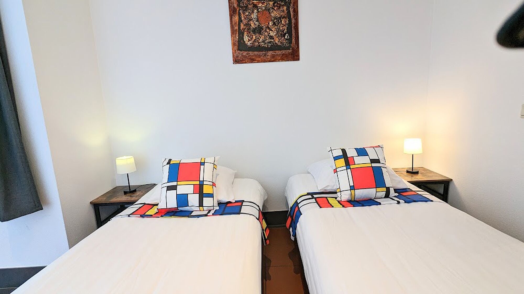 A hotel room with two single beds, each with a colorful geometric-patterned bedspread. There are bedside tables with lamps, a wall hanging above the beds, and a window on the left side.