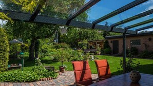 Un porche cubierto con una mesa de madera y dos sillas, rodeado de un jardín con diversas plantas y césped.
