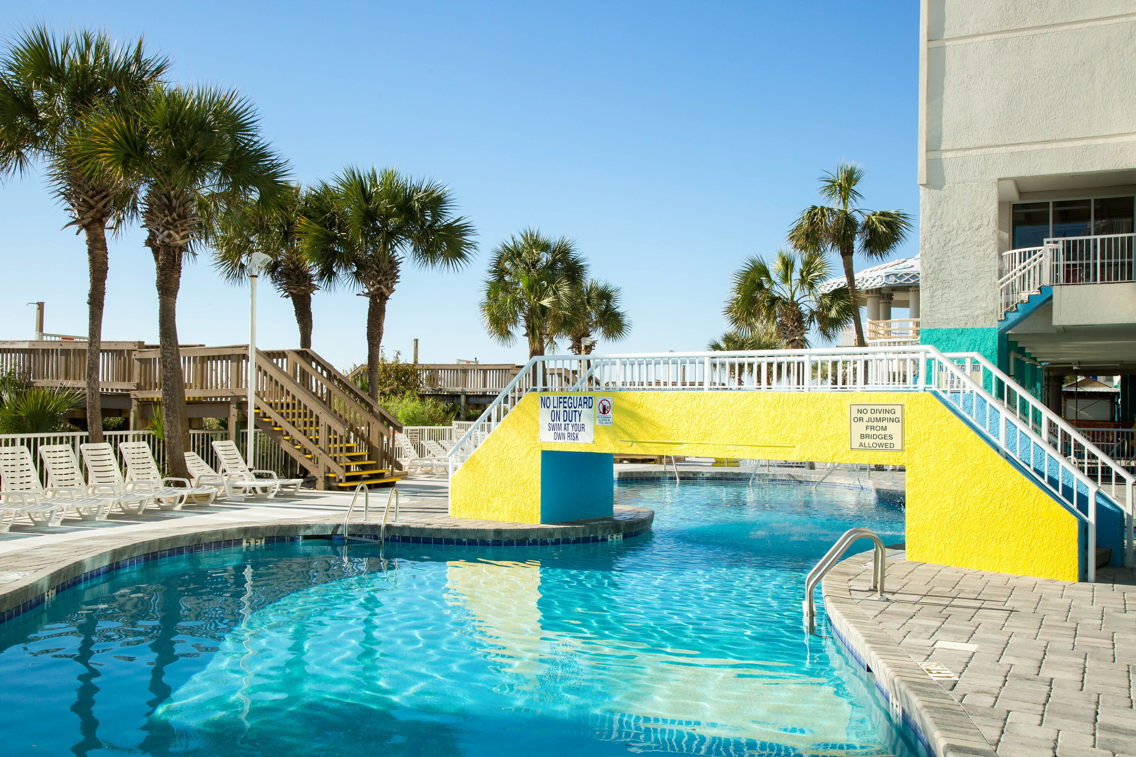 A swimming pool with a no lifeguard on duty sign, palm trees, and a staircase leading to a building.