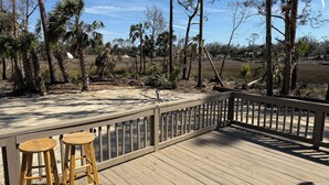 Una terraza de madera con dos taburetes de madera, con vistas a un paisaje natural con árboles y una zona arenosa.