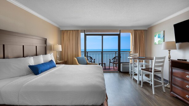 Ein Hotelzimmer mit einem großen Bett, einem Esstisch, Stühlen, einem Fernseher und einem Balkon mit Meerblick.