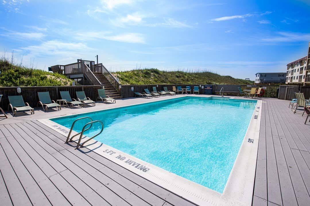 A rectangular swimming pool with a wooden deck surrounding it, equipped with lounge chairs and a safety sign.