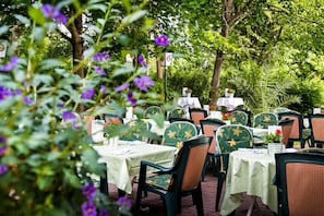Un área de comedor al aire libre con mesas y sillas, rodeada de abundante vegetación y árboles.