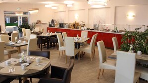 Area ristorazione di un hotel moderno, con tavoli rotondi, sedie beige e un bancone rosso con macchine per caffè.