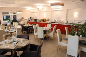 Ein moderner Hotel-Speisebereich mit runden Tischen, beigen Stühlen und einer roten Theke mit Kaffeemaschinen.