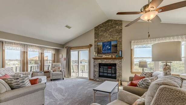 A spacious living room with a stone fireplace, comfortable seating, and large windows offering a view of the outdoors. - A Sitting Duck | 460 ft from the beach | Duck (Duck)