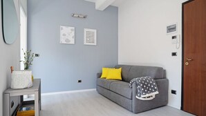 A modern living room with a grey sofa, yellow pillows, a white side table with a basket, and a grey blanket on the sofa. - GuestHost - The apartment is located in a quiet area, in a closed / private street overlooking the green and private gardens.It is located in a strategic position, a few steps from the historic center (the main road is located in absolute proximity to on (Bologna)