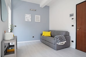 A modern living room with a grey sofa, yellow pillows, a white side table with a basket, and a grey blanket on the sofa.