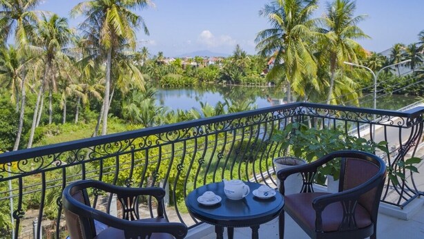 A balcony with a table and chairs, overlooking a body of water and palm trees. - hung hoang villa hoi an (Tp. Hội An)