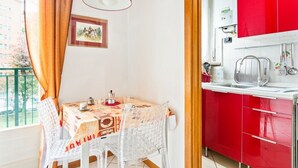 A dining area with a table set for two, a red kitchen with white cabinets, and a window with curtains. - GuestHost - NEW! Two-room apartment one step away from SAN SIRO STADIUM! (Milano)