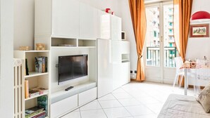 A modern living room with a flat-screen TV, white cabinetry, a dining area with a table and chairs, and a view of a building through a window with curtains. - GuestHost - NEW! Two-room apartment one step away from SAN SIRO STADIUM! (Milano)