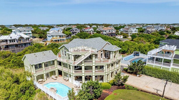 Une grande maison à plusieurs étages, dotée d’une piscine, entourée d’une végétation luxuriante et d’autres maisons.