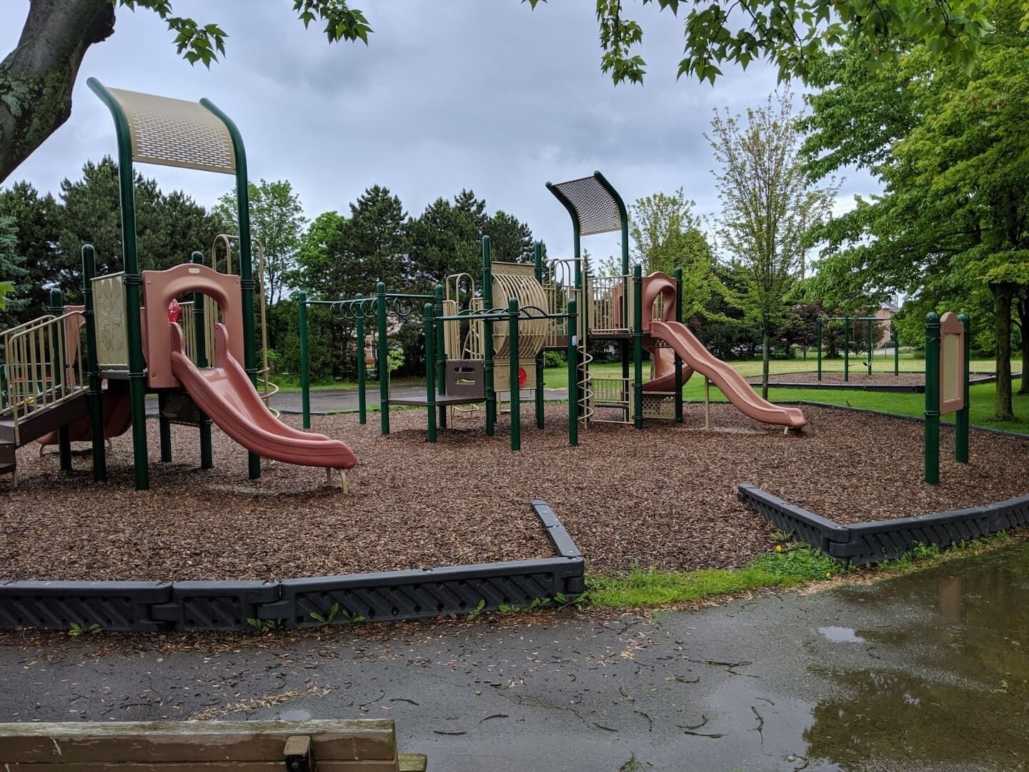 A playground with slides and climbing structures, surrounded by trees and a wet ground.