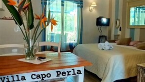 Una habitación acogedora con una mesa de madera que indica “Villa Capo Perla”, una cama, un televisor y un ventilador de techo.