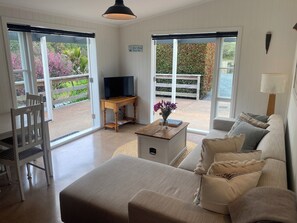 A spacious living room with a sofa, a coffee table, and a TV on a stand. There are large windows with a view of a garden and a wooden fence.
