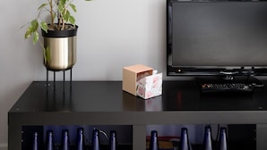 Uma lista emoldurada de destinos fictícios com códigos correspondentes, uma planta em vaso, uma televisão e um controle remoto sobre uma superfície preta.