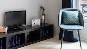 Uma sala de estar moderna com uma TV de tela plana sobre um suporte preto, uma poltrona azul com almofada estampada e um quadro na parede.