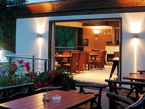 Ein Essbereich im Freien mit Tischen und Stühlen, ein Fernseher im Hintergrund und Blick ins Innere.