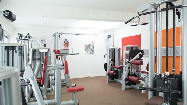 Ein Fitnessraum mit verschiedenen Trainingsgeräten, einer roten Bank und einer Wand mit einem Bild.