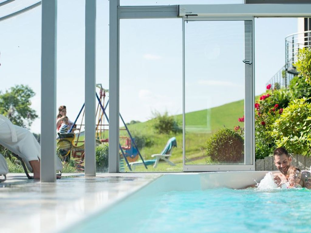 Una piscina interior con paredes de vidrio transparente, un tobogán y vistas a un paisaje verde.