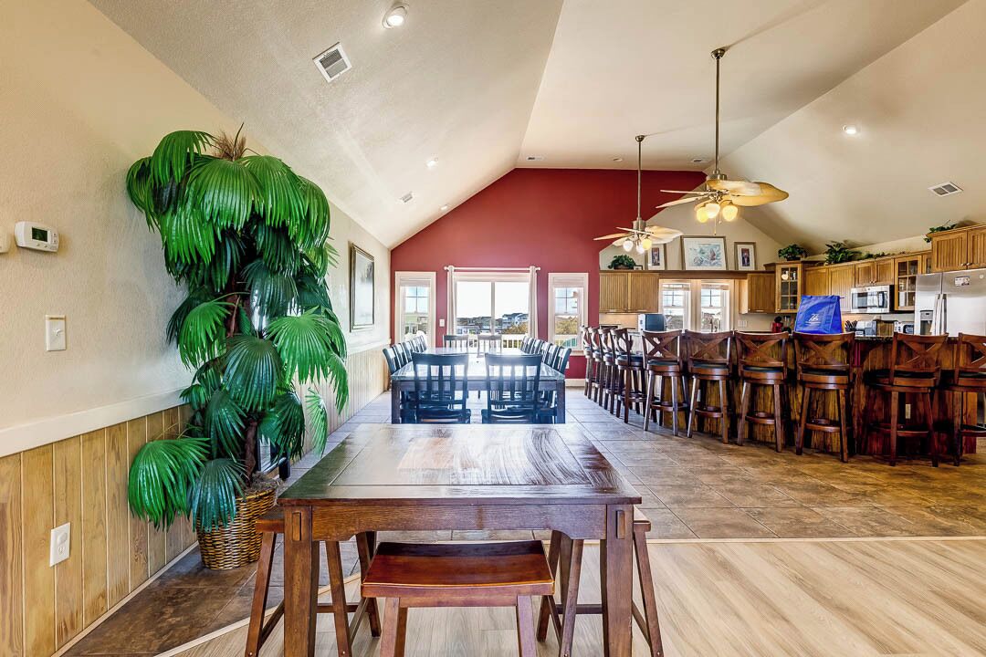 Ampia sala da pranzo con soffitto alto e pavimento in legno, caratterizzata da un grande tavolo circondato da sedie e un'alta pianta in vaso.