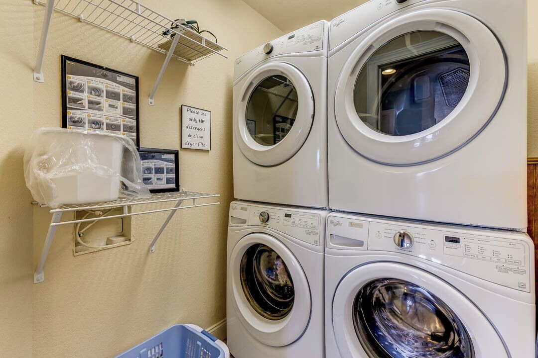 A laundry area with stacked washers and dryers, a shelf with laundry supplies, and a sign indicating the use of HE detergent and a clean drip filter.