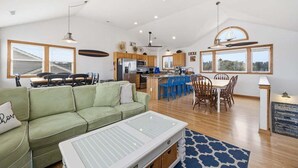 Uma sala ampla com um sofá verde, uma mesa de jantar com cadeiras azuis, uma cozinha com armários de madeira e janelas grandes que oferecem vista para o exterior.