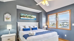 Quarto com uma cama grande, ventilador de teto, pintura de uma cena de praia e janela com vista para edifícios.