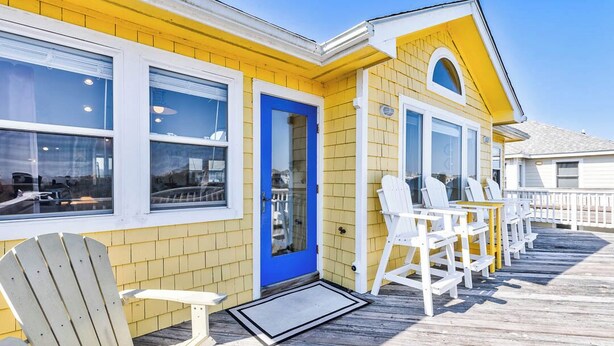 Um edifício amarelo com porta azul, janelas brancas e um deque de madeira com cadeiras brancas.