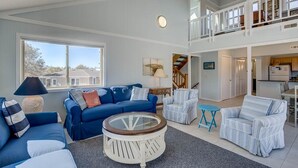 Un salon spacieux avec un canapé bleu, une table basse ronde et des chaises à rayures. Un escalier mène à l’étage supérieur.
