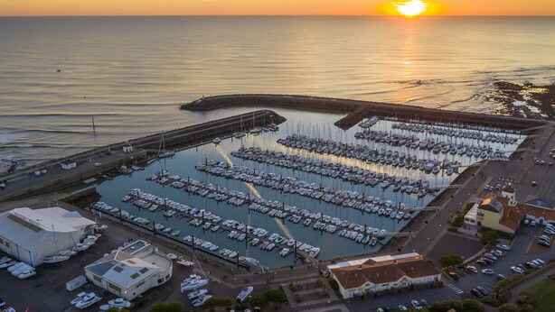 Ein Yachthafen mit vertäuten Booten, ein Gebäude mit flachem Dach und ein Sonnenuntergang über dem Wasser.