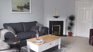 A living room with a grey sofa, a wooden coffee table with a book and a bottle, a fireplace with a decorative item, and a door.