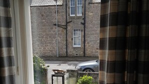 A room with a view of a brick building, a radiator, a chair with a white fur throw, and a candle in a glass holder on the windowsill.
