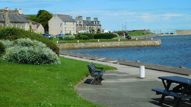 A waterfront promenade with benches, a stone wall, and buildings in the background.