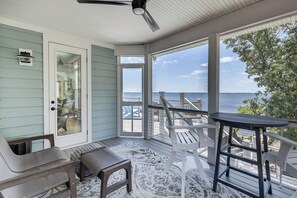 A covered porch with a view of the ocean, featuring a table and chairs, a bench, and a patterned rug.