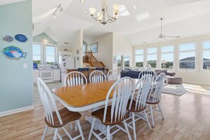 A spacious dining area with a wooden table and white chairs, a kitchen in the background, and large windows offering a view.