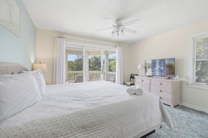 A bedroom with a large bed, a ceiling fan, a dresser, a television, and a balcony with a view of trees.