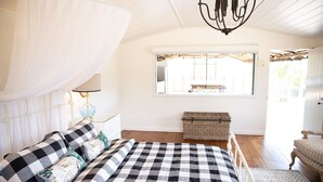 Un dormitorio con colcha a cuadros, dosel, lámpara de noche, un cofre de mimbre y vistas al exterior a través de una ventana.